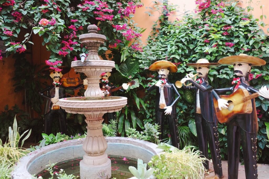 A courtyard scene featuring a decorative fountain surrounded by lush greenery and vibrant pink flowers. Around the fountain, there are statues of skeletal mariachi musicians wearing traditional outfits and sombreros, playing instruments like the trumpet and guitar. 