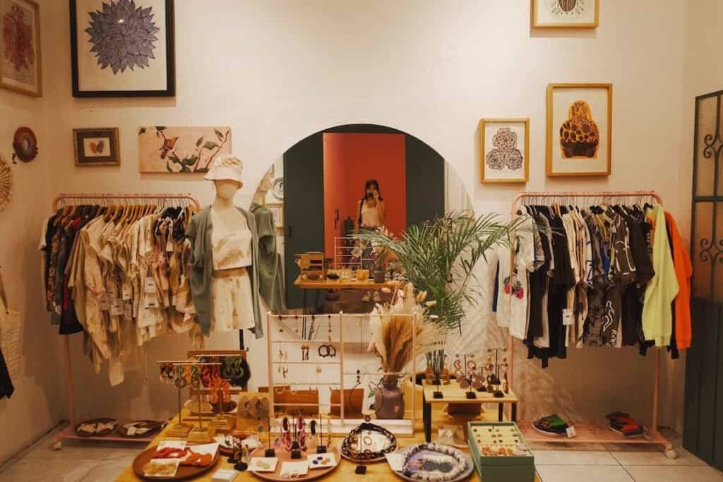 A boutique shop displays various handmade items, including jewelry, clothing, and accessories. A mannequin dressed in light, casual wear stands near racks of vibrant shirts and garments. The shop’s decor includes framed artwork on the walls, and a mirror in the center reflects the photographer taking the picture.