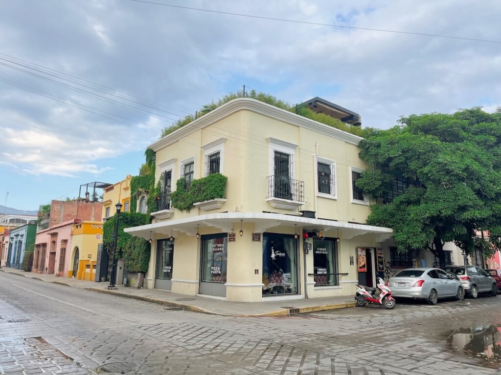 corner building in a colonial-style neighborhood, with light yellow walls and green plants climbing the exterior. The building has large windows with black iron bars and balconies, while the street features parked cars and a scooter. The road is made of cobblestones