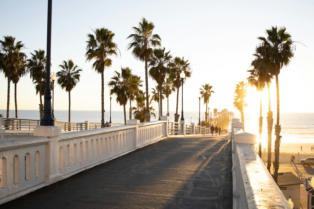 sunlit path leading to a pier, lined with tall palm trees on both sides. The warm glow of the setting sun reflects on the ocean and the white railings, while a few people walk along the path, enjoying the scenic beach view. The atmosphere is serene, with a mix of golden light and coastal vibes.