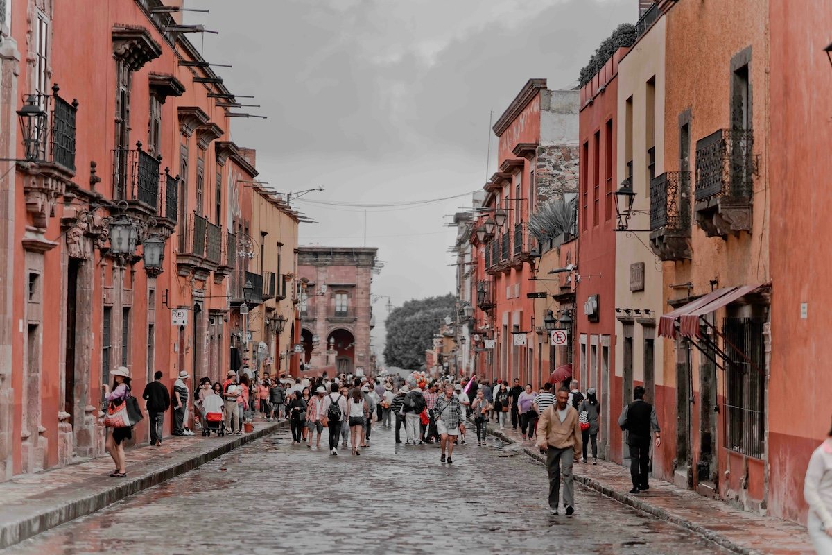 a lively cobblestone street in a historical city, with people walking amidst colorful, colonial-style buildings. The walls of the buildings are painted in warm shades of red, orange, and yellow, and ornate iron balconies and vintage street lamps line the street. The overcast sky adds a soft, diffused light to the scene, enhancing the charm of this bustling town.