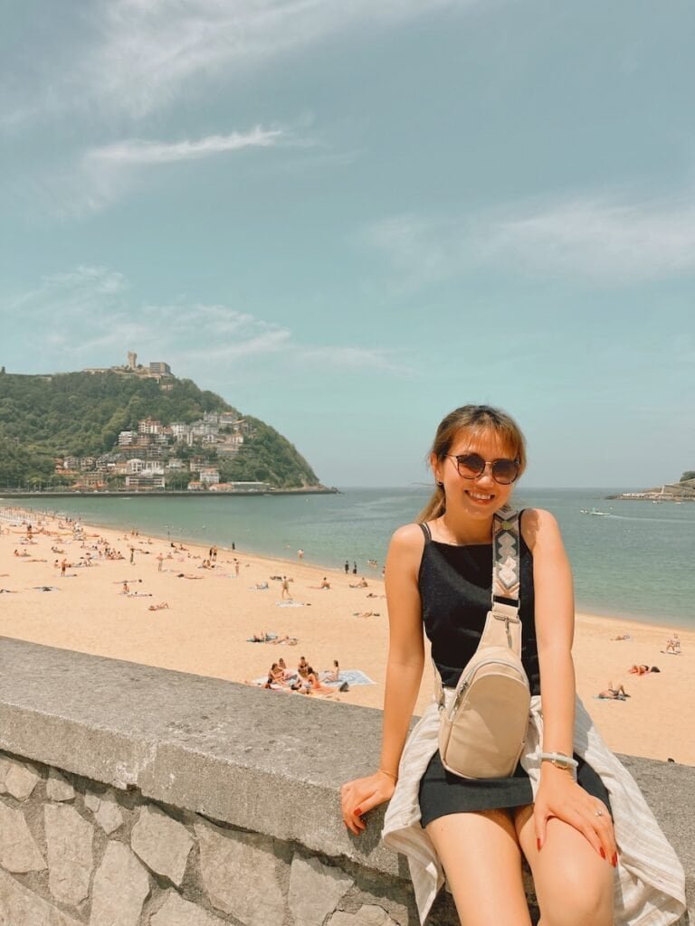 This image shows a smiling woman sitting on a stone wall overlooking a beach on a sunny day. Behind her, there are people relaxing on the sandy beach with a scenic hillside and water in the background. The woman is wearing sunglasses, a black sleeveless dress, and a crossbody bag, with a light jacket tied around her waist. The overall setting appears peaceful and summery.