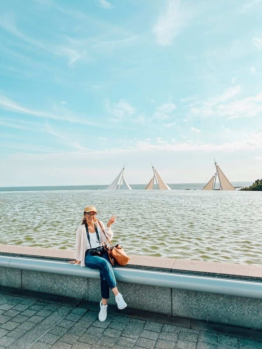 This image features a woman sitting on a ledge near a waterfront, smiling and holding up a peace sign. She is wearing a light sweater, jeans, white sneakers, and a baseball cap, with a large brown bag beside her and a camera around her neck. In the background, three sailboats are partially submerged in the water, with only their sails visible above the surface under a bright blue sky.