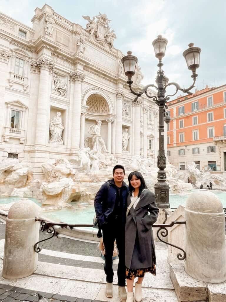 A couple stands in front of the Trevi Fountain in Rome, smiling at the camera. The ornate details of the baroque architecture and statues behind them are captured beautifully, while the light blue water of the fountain creates a calm atmosphere. They are dressed warmly, suggesting a cooler day.