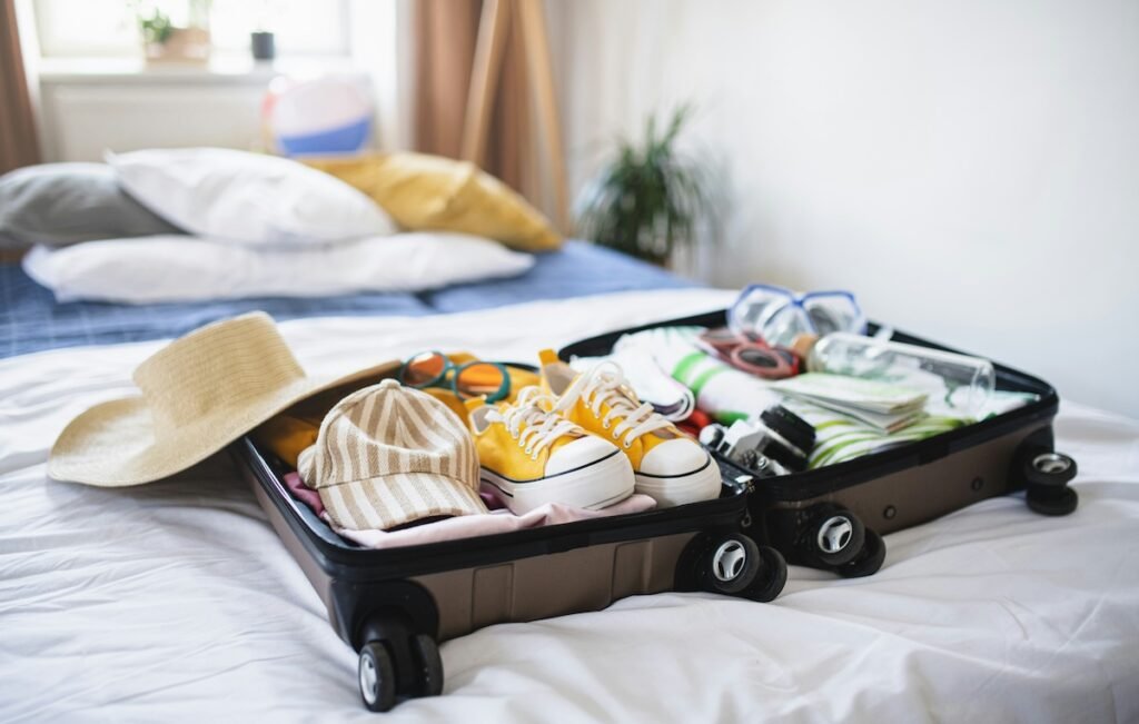 A suitcase lies open on a bed, filled with travel essentials like a pair of yellow sneakers, a striped cap, sunglasses, and snorkeling gear. A straw hat rests on the side of the suitcase, with neatly folded clothes visible underneath the accessories. In the background, soft pillows and a blurred bedroom