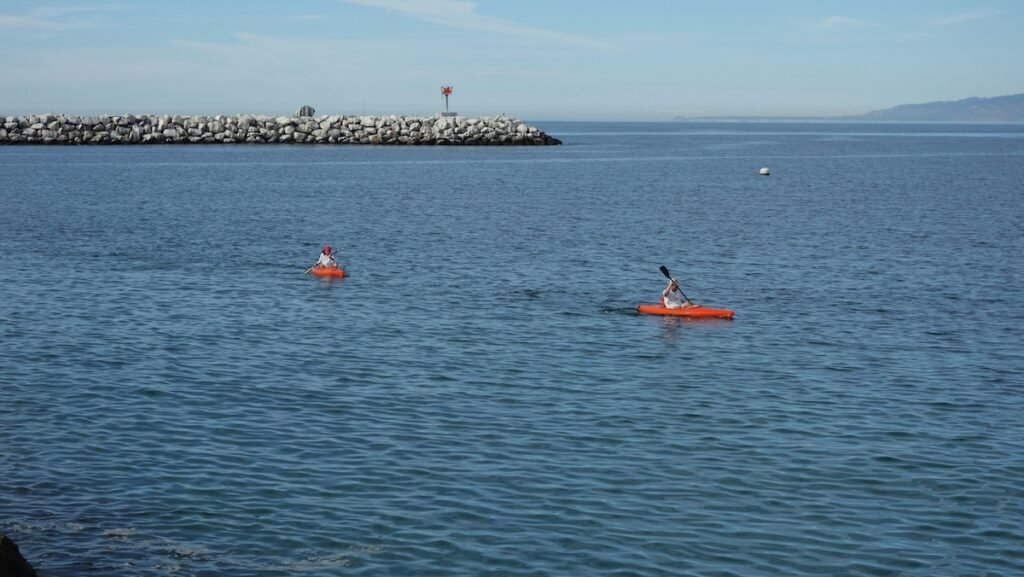 Two kayakers paddle across the calm blue waters near a rocky breakwater at Playa Del Rey. The kayaks are bright orange, contrasting against the serene ocean. A distant mountain range and a small red navigation marker are visible in the background, enhancing the tranquil coastal scene.