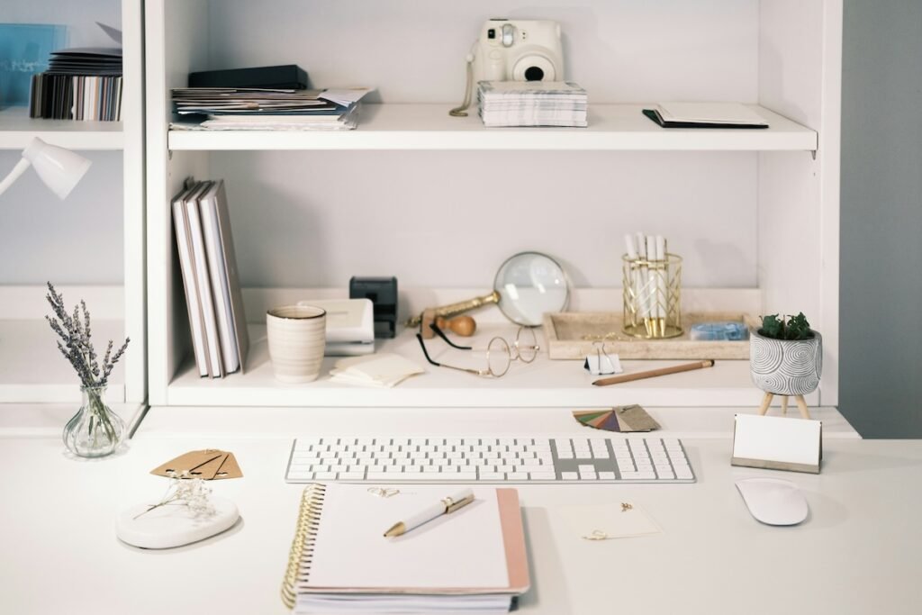 A minimalist white desk setup with a keyboard, mouse, and a spiral notebook with a pen resting on it. The shelves above hold neatly arranged items such as a Polaroid camera, books, a small plant, and various stationery like a magnifying glass and glasses. A small vase with dried lavender adds a touch of natural decor to the otherwise organized workspace.