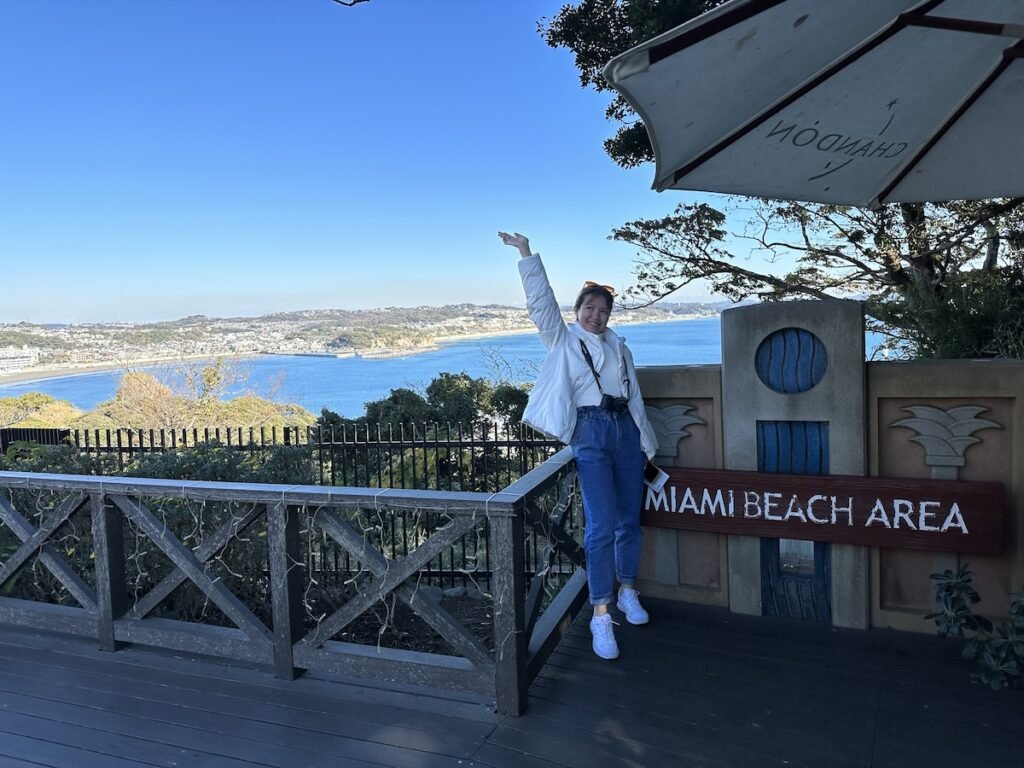 Miami Beach sign at the observation deck overlooking the water and mainland. A girl with jeans and white puffer jacket is standing next to the sign with her right arm up in the air