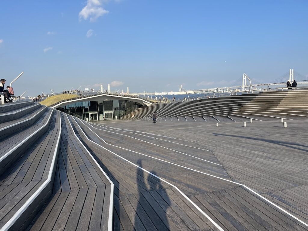  sunny day at Osanbashi Pier Terminal in Yokohama, showcasing its distinctive architectural design with sweeping wooden decks and modern, angular lines. People can be seen strolling and relaxing, while in the background, the Yokohama Bay Bridge adds to the scenic coastal view under a clear blue sky. The pier blends urban sophistication with a serene waterfront vibe.