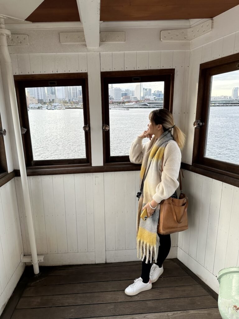 woman in winter clothes standing in front of a ship cabin with windows looking to the ocean