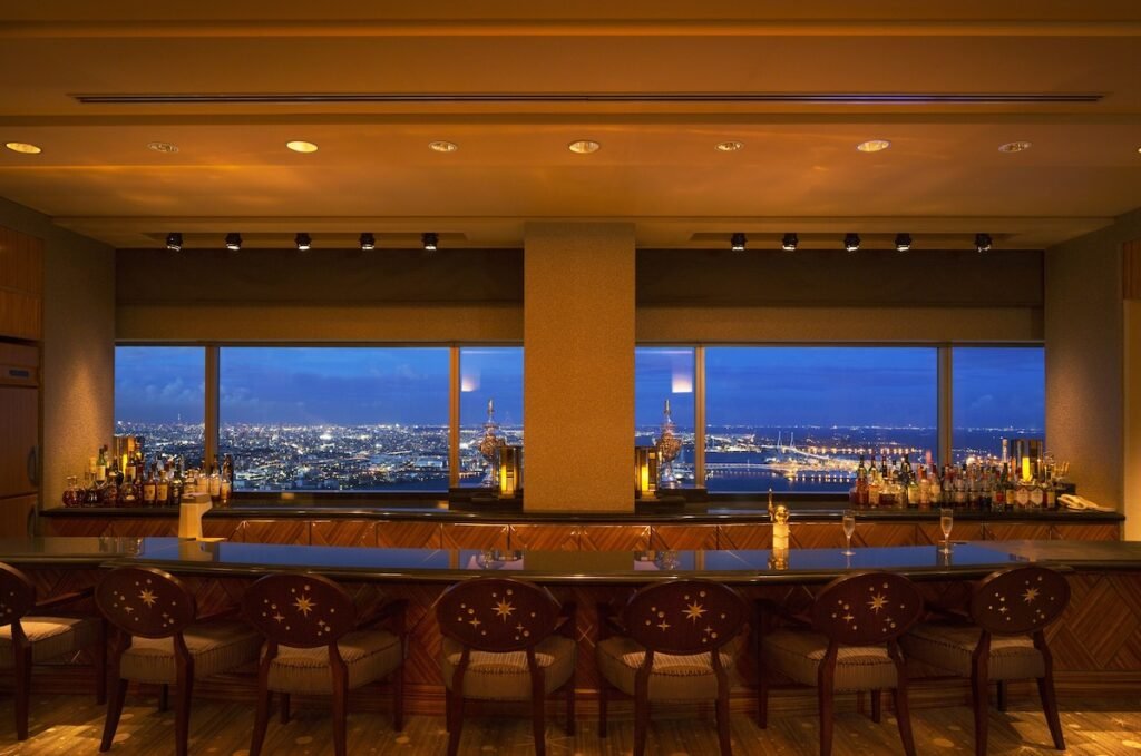 An elegant rooftop bar with a luxurious wooden counter and star-patterned chairs, offering a panoramic view of a brightly lit cityscape at dusk. The large windows frame the sprawling urban skyline, adorned with twinkling lights and a serene harbor in the distance. The bar is warmly lit, creating an inviting atmosphere perfect for evening relaxation.