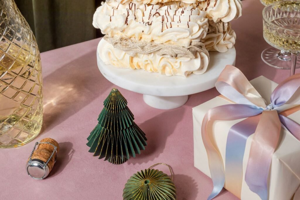 A festive holiday table setup featuring a marble cake stand holding a pavlova dessert with whipped cream and chocolate drizzle, surrounded by Christmas decorations including miniature green paper trees and a gift box wrapped in pastel ribbon. A crystal decanter and champagne glasses add elegance to the scene, evoking a celebratory atmosphere perfect for the winter season.