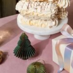 A festive holiday table setup featuring a marble cake stand holding a pavlova dessert with whipped cream and chocolate drizzle, surrounded by Christmas decorations including miniature green paper trees and a gift box wrapped in pastel ribbon. A crystal decanter and champagne glasses add elegance to the scene, evoking a celebratory atmosphere perfect for the winter season.