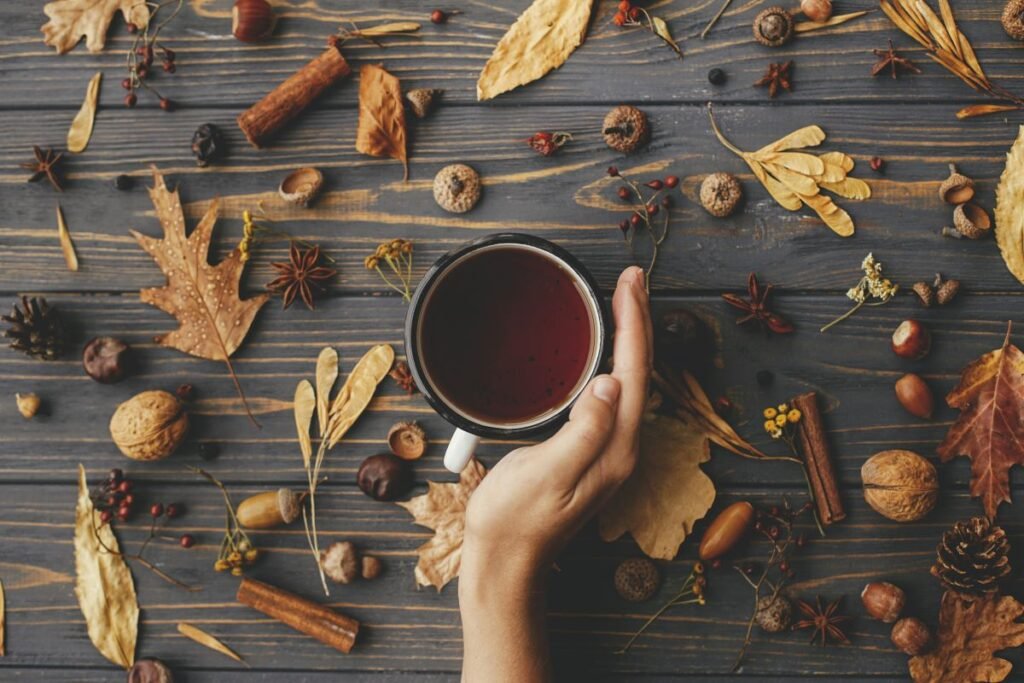 A cozy autumn-themed flat lay featuring a hand holding a mug of tea, surrounded by fall elements such as dried leaves, cinnamon sticks, acorns, walnuts, star anise, and pinecones. The wooden table adds a rustic feel, while the warm tones evoke a seasonal, comforting atmosphere. Perfect for illustrating fall beverages or seasonal mood boards.