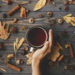 A cozy autumn-themed flat lay featuring a hand holding a mug of tea, surrounded by fall elements such as dried leaves, cinnamon sticks, acorns, walnuts, star anise, and pinecones. The wooden table adds a rustic feel, while the warm tones evoke a seasonal, comforting atmosphere. Perfect for illustrating fall beverages or seasonal mood boards.