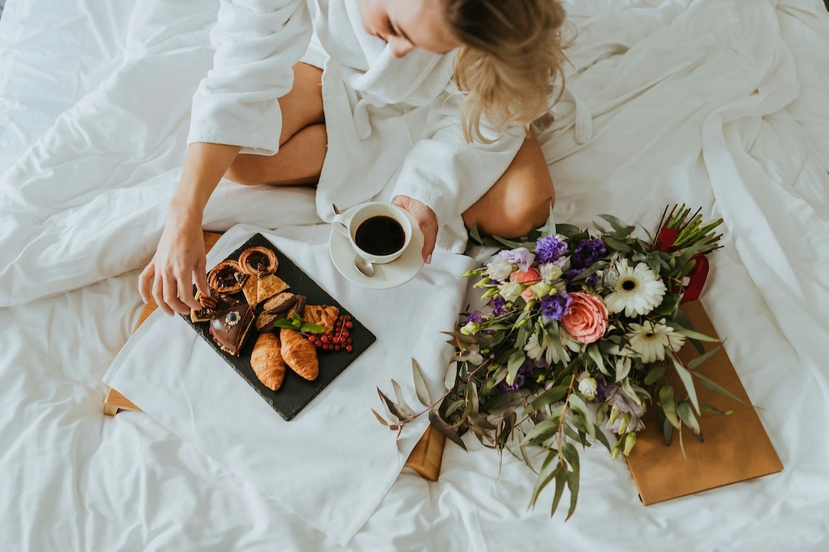 A serene morning scene featuring a woman in a white robe enjoying breakfast in bed. The tray includes an assortment of pastries, chocolates, and berries, paired with a cup of coffee. Beside her lies a colorful bouquet of flowers, adding a touch of elegance and warmth to the cozy setting.