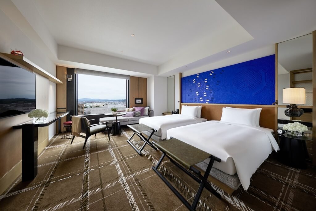 This image showcases a modern hotel room with two twin beds, a large window with a city view, and contemporary decor. The room features a striking blue accent wall with an artistic design, a cozy seating area near the window, and elegant furnishings including a glass table, a sleek TV setup, and stylish lighting. The neutral tones and soft lighting create a luxurious yet inviting atmosphere.