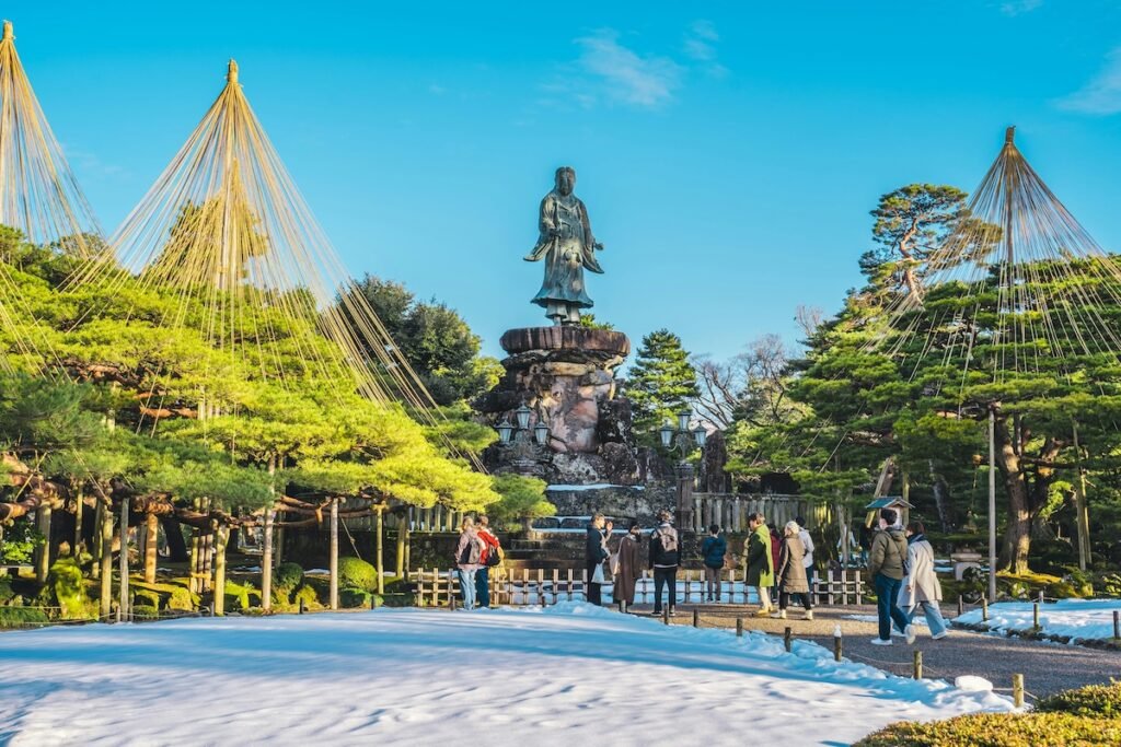 beautiful snowy day at the kenroku-en garden in kanazawa. there's people strolling in the park among green trees. There's a copper statue in the background