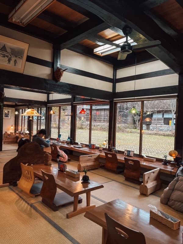 cozy interior of a traditional coffee shop with wooden accents on the roof and low seating wooden tables and chairs. The coffee shop has large glass windows facing the shirakawago village