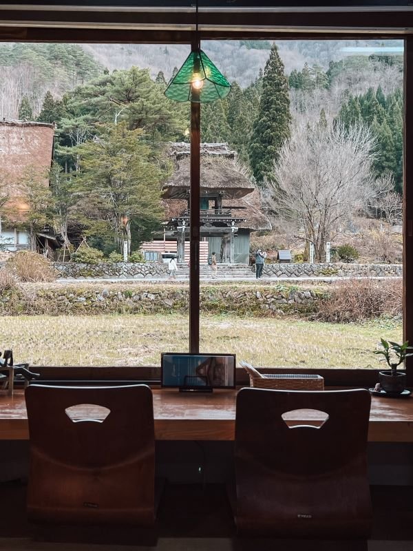 image of a low wooden table and wooden chairs facing large glass window with a view of thatched houses in shirakawago.