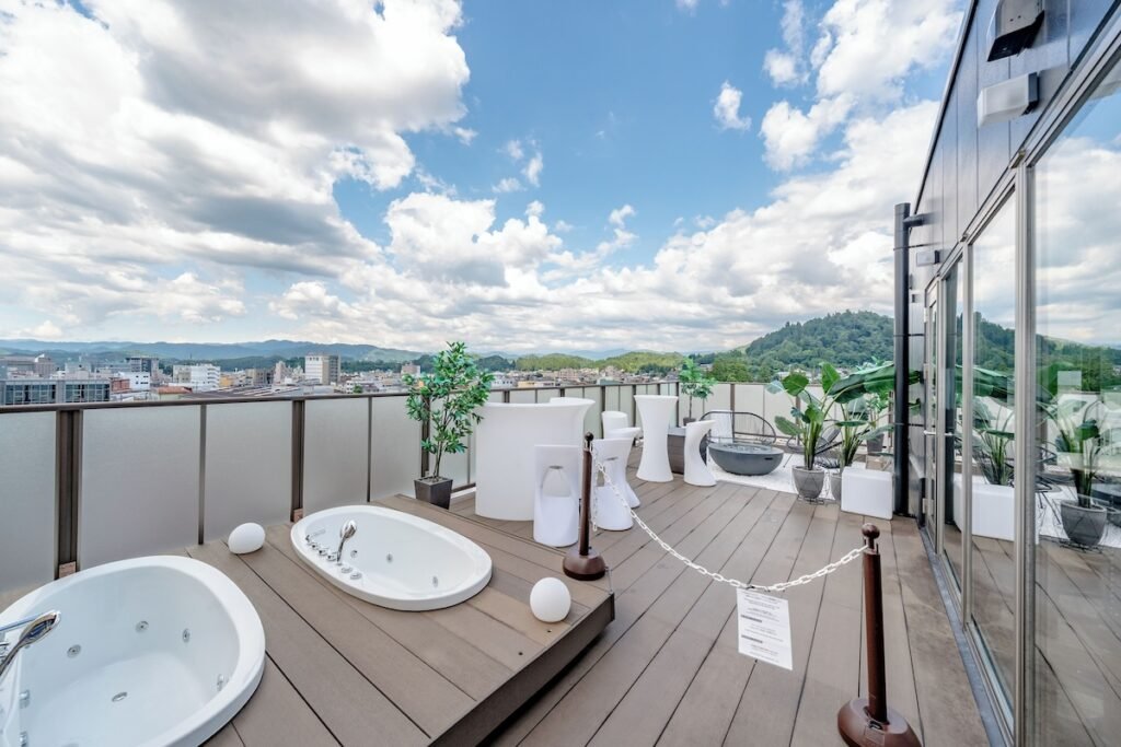 This image showcases a stylish rooftop terrace with two outdoor jacuzzis, modern white furniture, and lush greenery, offering a panoramic city and mountain view. The wooden deck creates a warm, inviting atmosphere, while the sleek decor, including tall cocktail tables and contemporary seating, suggests a luxurious space for relaxation or events. The bright blue sky with scattered clouds enhances the open and airy feel of the setting.